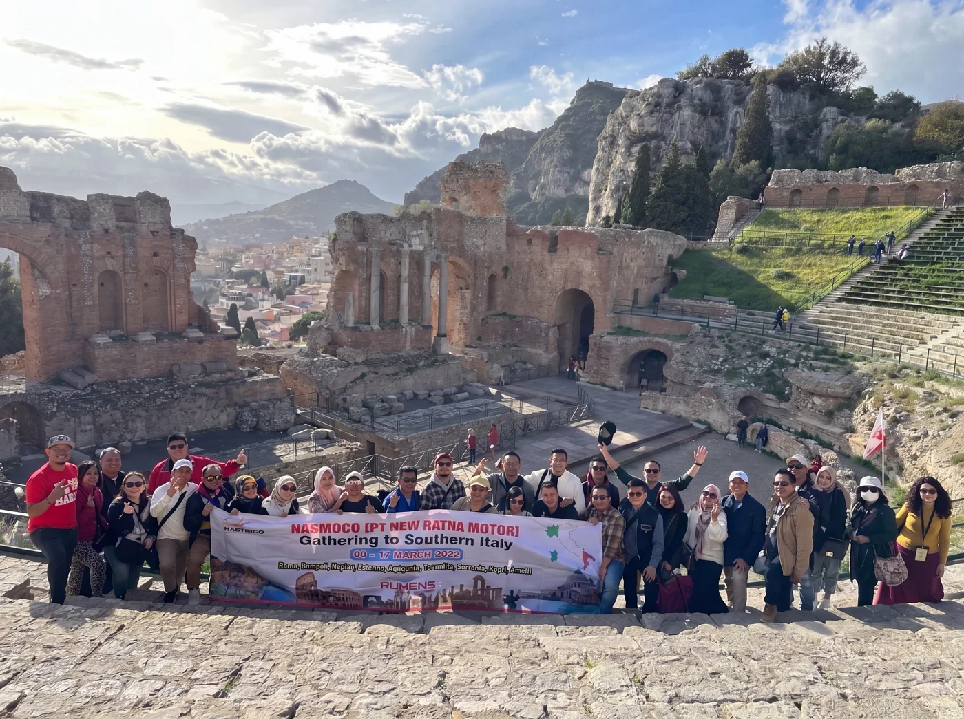 Peserta perjalanan Ruhens berfoto bersama spanduk grup di depan teater batu bersejarah dan perbukitan di Italia.