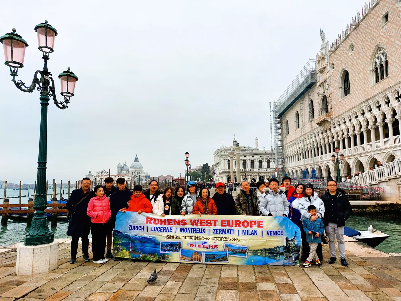 Peserta perjalanan berfoto dengan spanduk Ruhens di tepi perairan Venesia, dengan bangunan bersejarah di belakangnya.