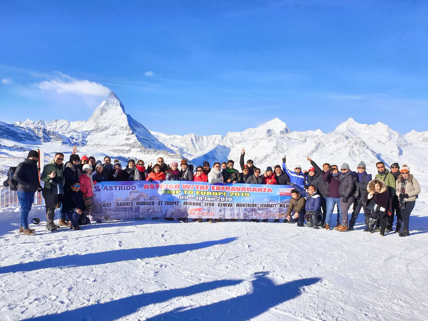 Peserta perjalanan Ruhens dengan spanduk grup di lanskap bersalju Zermatt, dengan puncak gunung di belakangnya.
