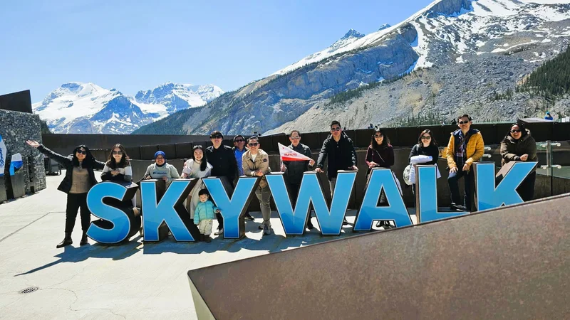 Grup perjalanan berfoto di belakang tulisan Skywalk dengan pegunungan bersalju di belakangnya.