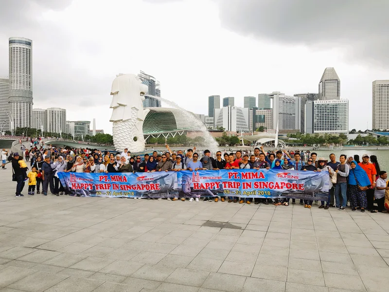 Grup MIMA dengan spanduk di tepi perairan Singapura, dengan Merlion dan gedung kota di belakangnya.
