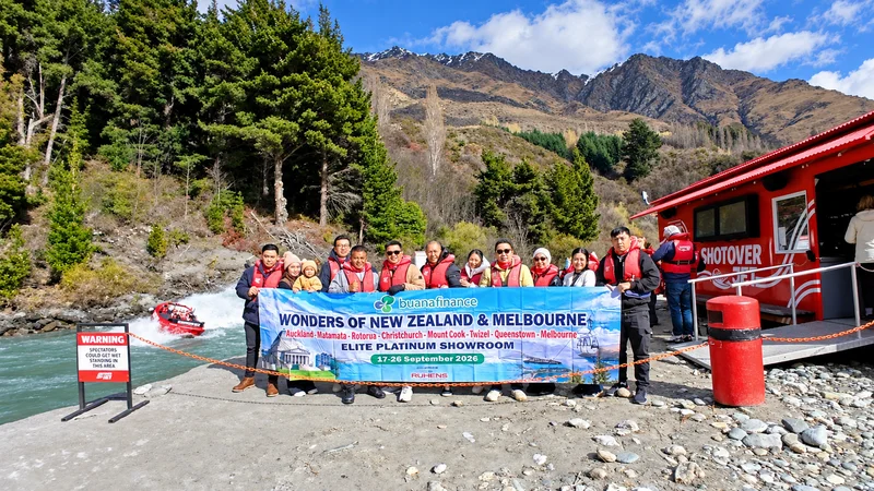 Grup perjalanan berfoto dengan spanduk Buana Finance di tepi sungai, dengan kapal merah dan pegunungan di belakangnya.