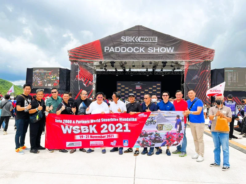Grup perjalanan dengan spanduk Auto 2000 WSBK 2021 di depan panggung Paddock Show Mandalika.