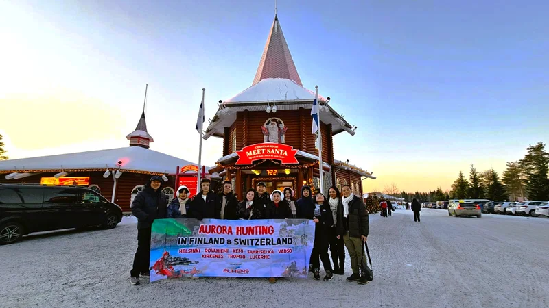 Grup perjalanan dengan spanduk Aurora Hunting berfoto di depan bangunan kayu di lanskap bersalju.
